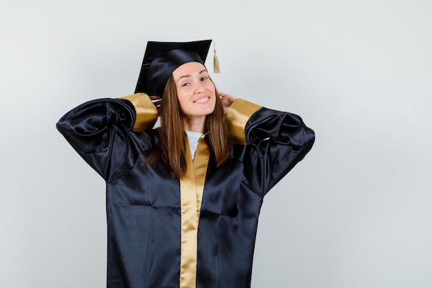 Foto gratuita giovane laureato femminile che tiene le mani dietro la testa in abito accademico e guardando allegro, vista frontale.