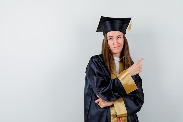 Молодая женщина-выпускница указывает на верхний правый угол в академической одежде и выглядит недовольной. передний план.