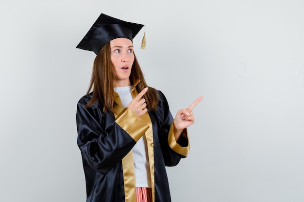 Young female graduate pointing at upper right corner in academic dress and looking puzzled , front view.