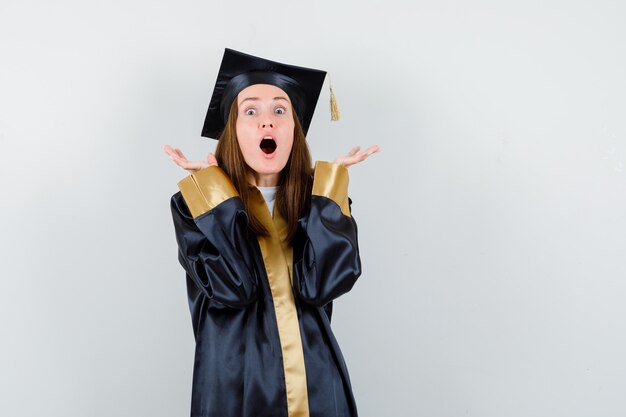 Young female graduate raising hands in academic dress and looking amazed. front view.