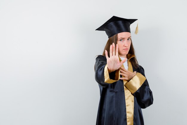 Young female graduate showing stop gesture in academic dress and looking serious , front view.