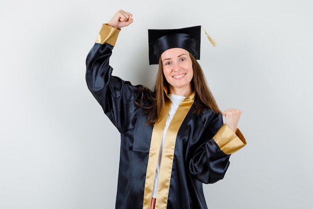 Молодая женщина-выпускница показывает жест победителя в академической одежде и выглядит удачливым, вид спереди.