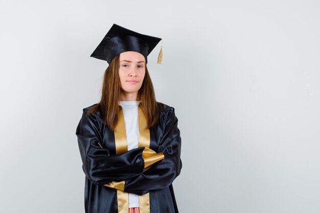 Young female graduate standing with crossed arms in academic dress and looking confident. front view.