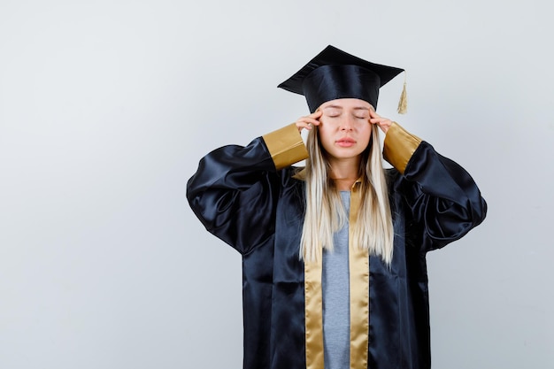 Young female in graduate uniform keeping fingers on her temples and looking tired