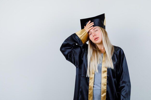 Young female in graduate uniform keeping hand on head and looking distressed