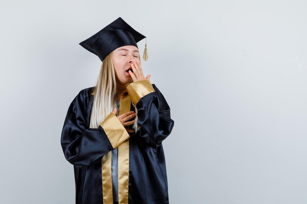 Молодая женщина в униформе выпускника держит руку на рту, зевая и выглядя сонной
