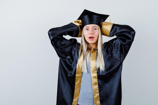 Young female in graduate uniform keeping hands behind head and looking amazed