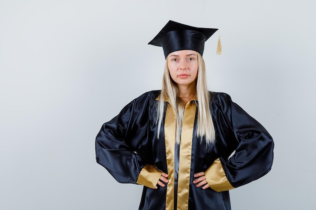 腰に手を保ち、自信を持って見える大学院の制服を着た若い女性