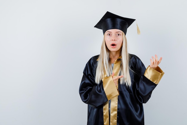 Free photo young female in graduate uniform pointing aside and looking surprised