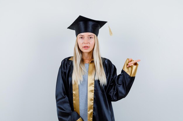 Молодая женщина в униформе выпускника, указывающая вправо и выглядящая разумной