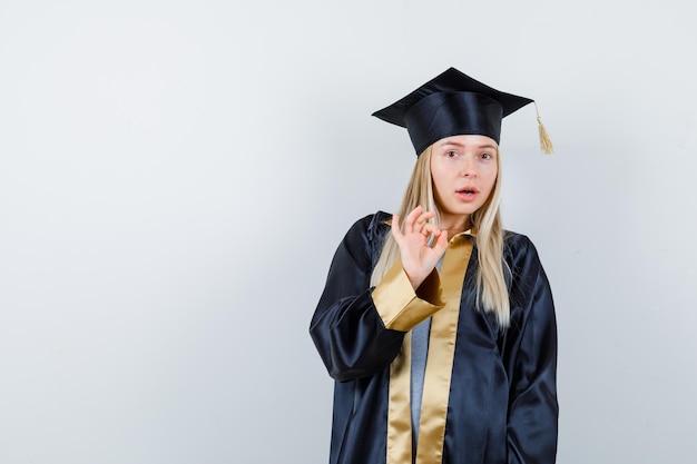 Young female in graduate uniform showing ok gesture and looking puzzled,