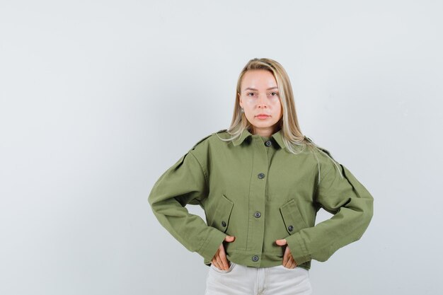 Молодая женщина в зеленой куртке, джинсах, взявшись за руки в кармане и серьезно, вид спереди.