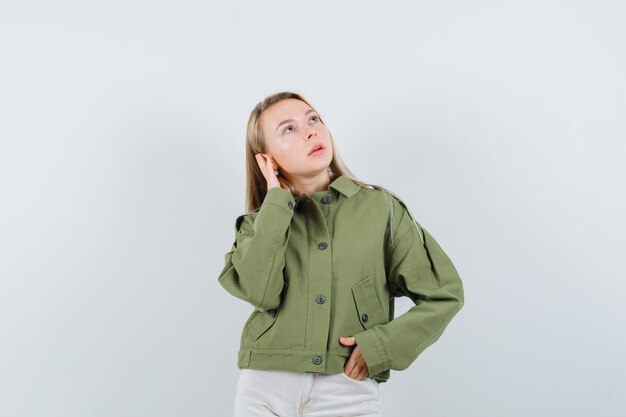 Молодая женщина в зеленой куртке, джинсах позирует с рукой на ухе и выглядит впечатляюще, вид спереди.