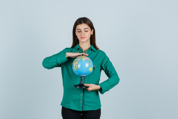 Foto gratuita giovane donna in camicia verde guardando il globo terrestre e guardando attento, vista frontale.