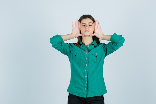 Giovane donna in camicia verde in posa con le mani sui lati del viso e guardando rilassato, vista frontale.