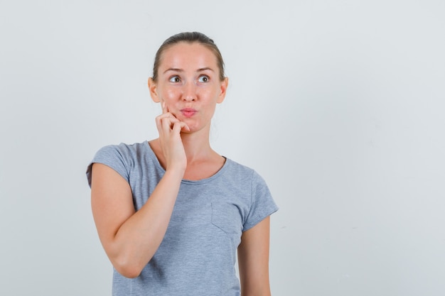 Young female in grey t-shirt holding finger on cheek and looking pensive , front view.