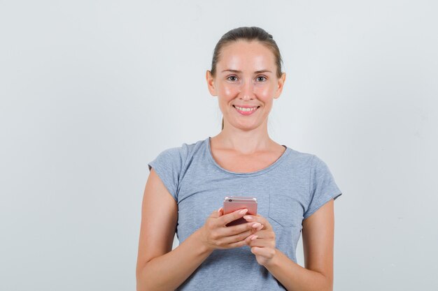 Молодая женщина в серой футболке держит мобильный телефон и выглядит веселым, вид спереди.