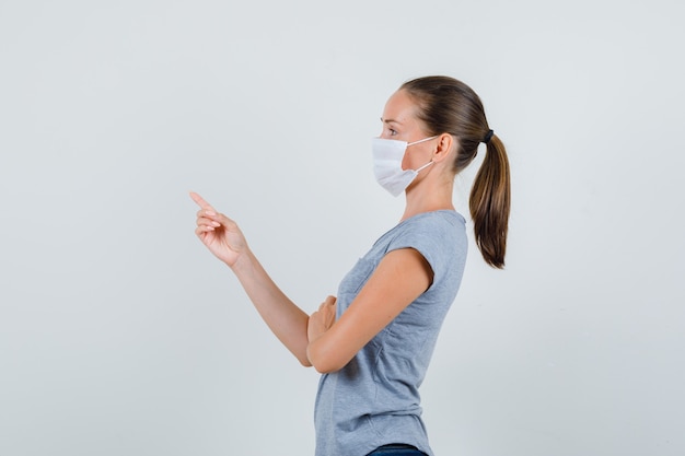 Young female in grey t-shirt, jeans, mask pointing at something away .