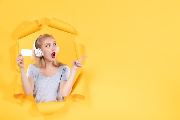 노란색 벽에 신용카드가 있는 헤드폰을 끼고 있는 젊은 여성