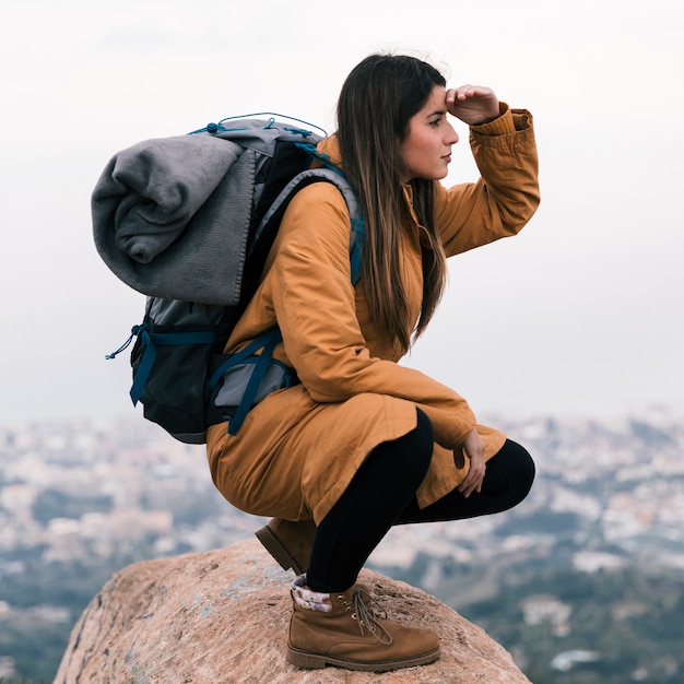 Молодая женщина hiker, сидя на вершине скалы с ее рюкзаком, экранируя глаза