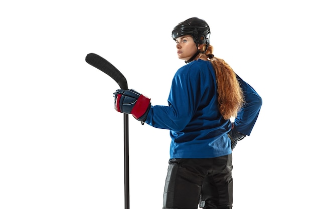 Young female hockey player with the stick on ice court and white wall. Sportswoman wearing equipment and helmet posing. Concept of sport, healthy lifestyle, motion, action, human emotions.