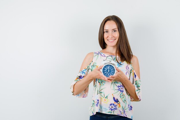 Young female holding alarm clock in shirt, jeans and looking cheery , front view.