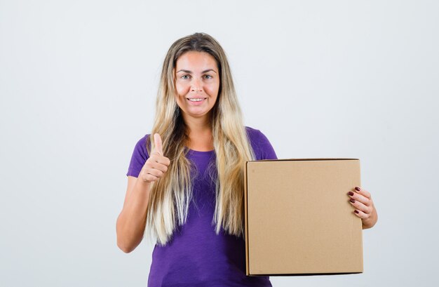 紫色のTシャツに親指を表示し、嬉しそうに見える、正面図の若い女性の保持ボックス。