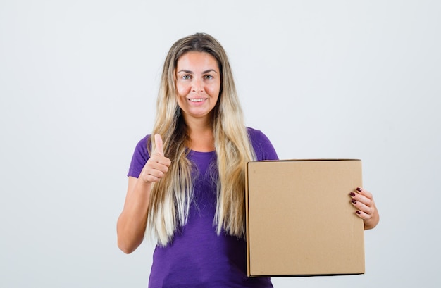 Young female holding box while showing thumb up in violet t-shirt and looking glad , front view.