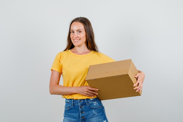 Free photo young female holding cardboard box and smiling in t-shirt, shorts front view.