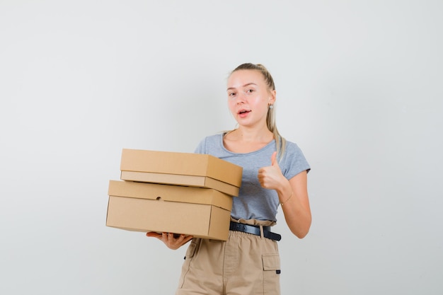 Free photo young female holding cardboard boxes, showing thumb up in t-shirt, pants and looking cheery. front view.