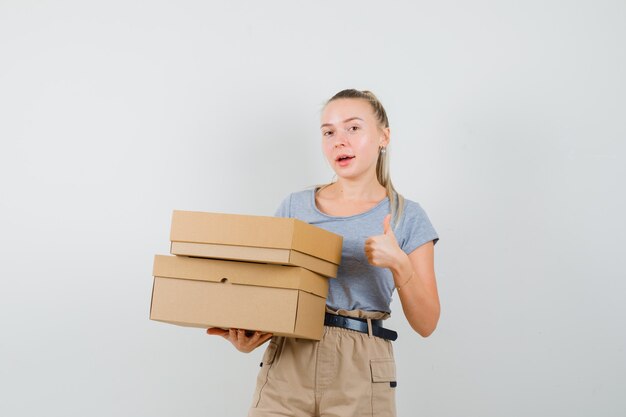 Молодая женщина держит картонные коробки, показывает палец вверх в футболке, штанах и выглядит весело. передний план.