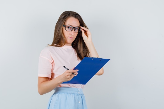 Young female holding clipboard and pen in t-shirt, skirt and looking careful. front view.