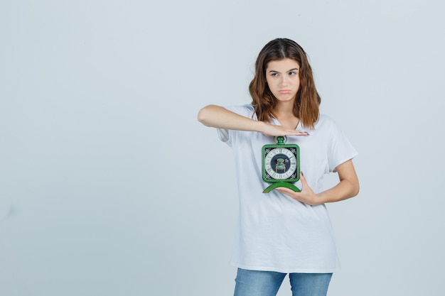 Young female holding clock in white t-shirt, jeans and looking hesitant , front view.