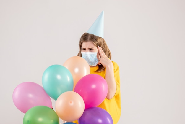 Young female holding colorful balloons in mask on white