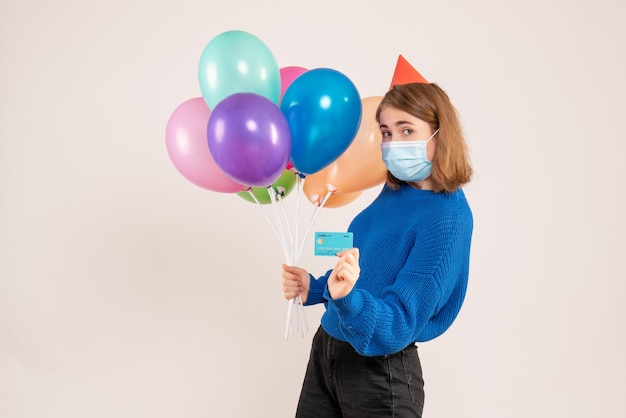 young female holding colorful balloons in mask with bank card on white