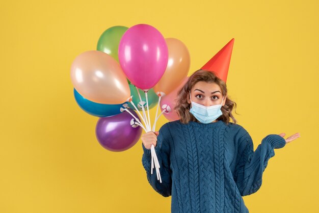 young female holding colorful balloons in mask on yellow