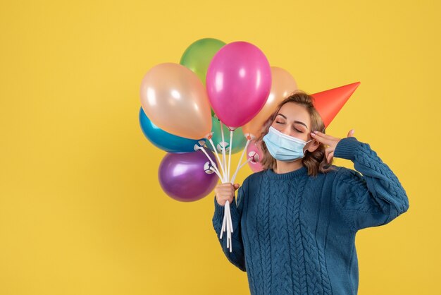 young female holding colorful balloons in mask on yellow