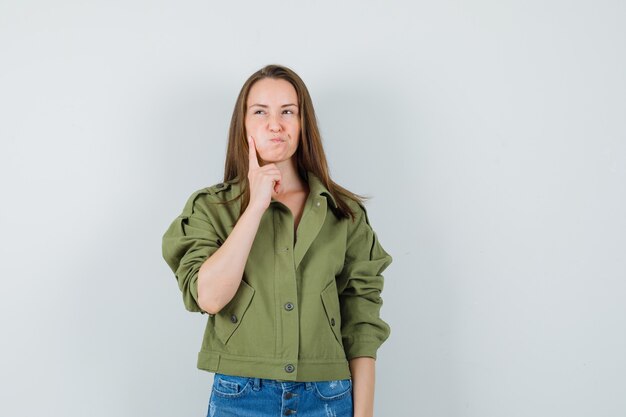 Young female holding finger on blown cheek in jacket, shorts and looking hesitant , front view.