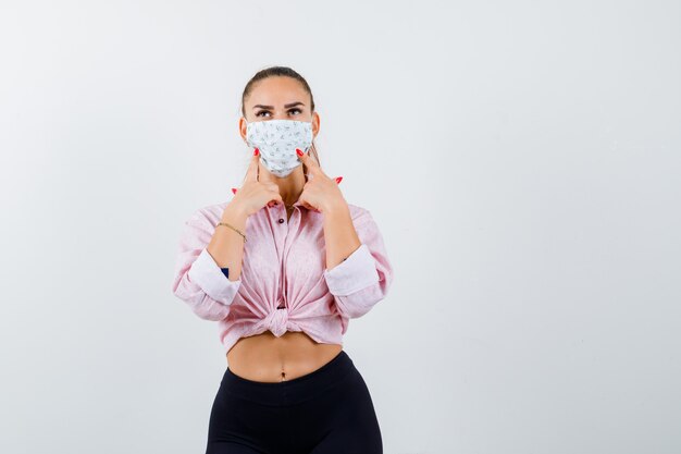 Free photo young female holding fingers on cheeks in shirt, pants, medical mask and looking cute , front view.