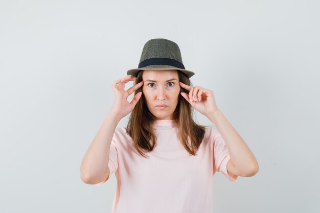 Young female holding fingers on temples in pink t-shirt, hat and looking serious. front view.