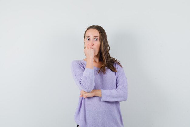 Young female holding fist on her mouth in lilac blouse and looking anxious. front view. space for text