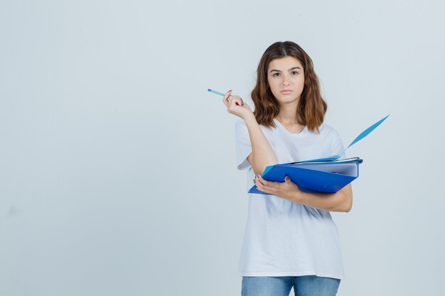 Молодая женщина, держащая папки и ручку в белой футболке, джинсах и задумчивая, вид спереди.