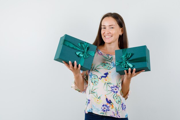 Young female holding gift boxes in shirt, jeans and looking cheerful , front view.