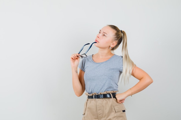 Young female holding glasses while looking up in t-shirt, pants and looking thoughtful. front view.