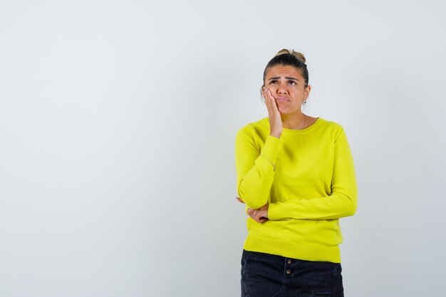 Young female holding hand on cheek in sweater, denim skirt and looking pensive