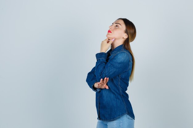 Young female holding hand under chin in denim shirt and jeans and looking thoughtful