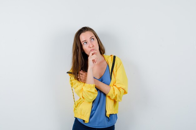 Молодая женщина держит руку на подбородке в футболке, куртке и смотрит задумчиво, вид спереди.