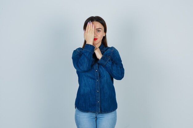 Young female holding hand on eye in denim shirt and jeans and looking serious