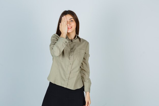 Young female holding hand on eye in shirt, skirt and looking merry