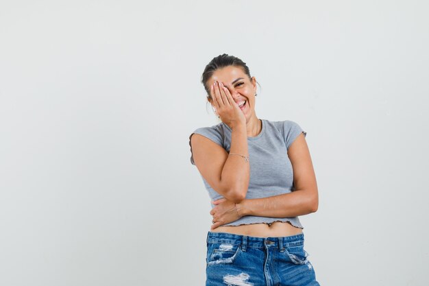 Young female holding hand on face in t-shirt, shorts and looking cheerful